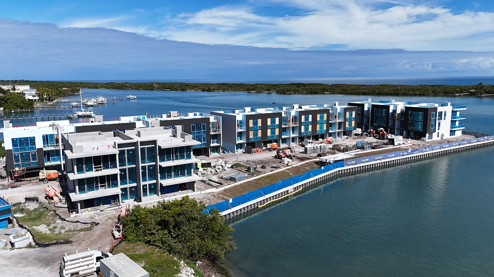 Aerial view of modern luxury waterfront townhomes under construction beside turquoise water and marina.